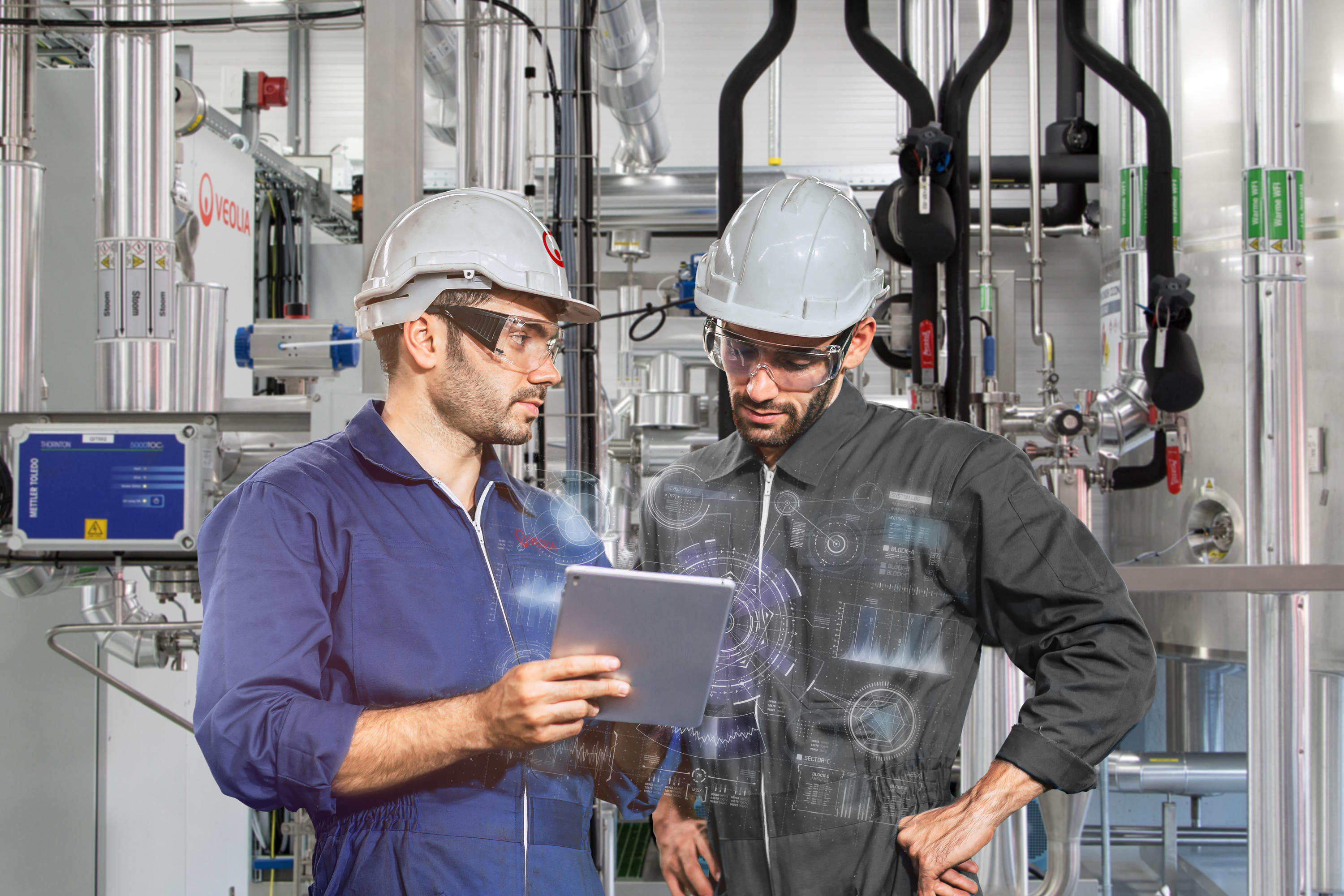 Two man wearing safety helmets, glasses and jumpsuits, holding a tablet, talking, working in a factory.