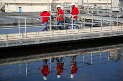 Three men from the staff, wearing uniforms and helmets, walking next to each other in a wastewater treatment plant, near water reservoir.
